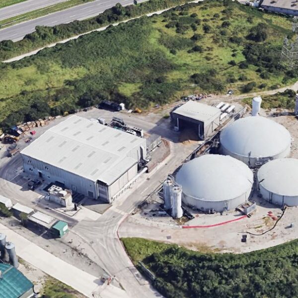 Aerial view of an industrial biogas plant surrounded by greenery. Three large, round silos are adjacent to a rectangular building, with roads nearby.